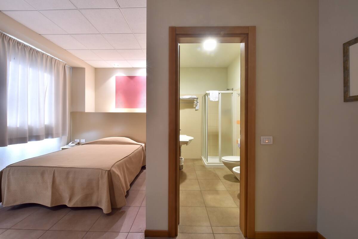 A hotel room with a bed, a bedside table, a window with curtains, and a bathroom visible through an open door.