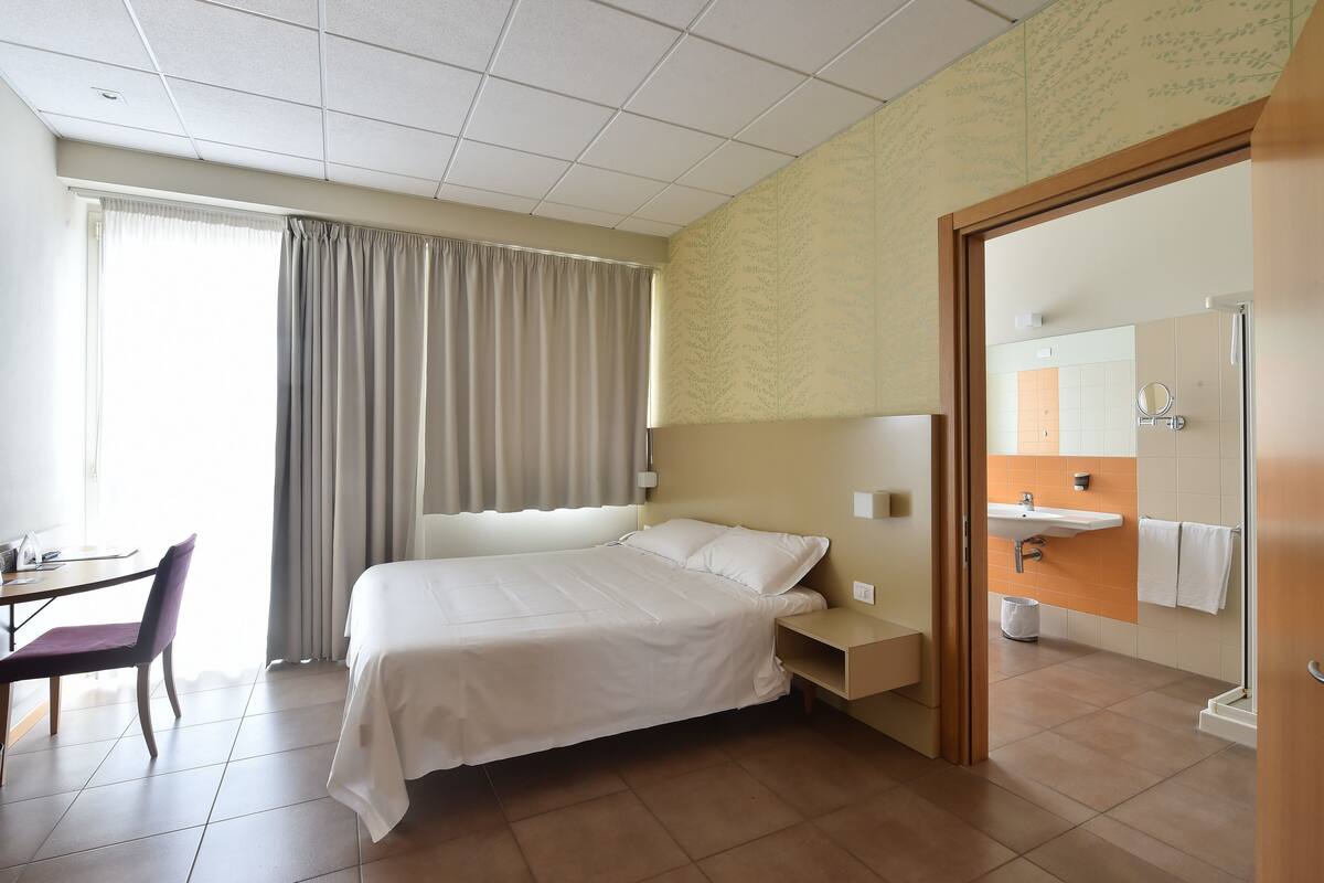A hotel room with a bed, a desk, a chair, a nightstand, a bathroom with a sink and mirror, and a window with curtains.