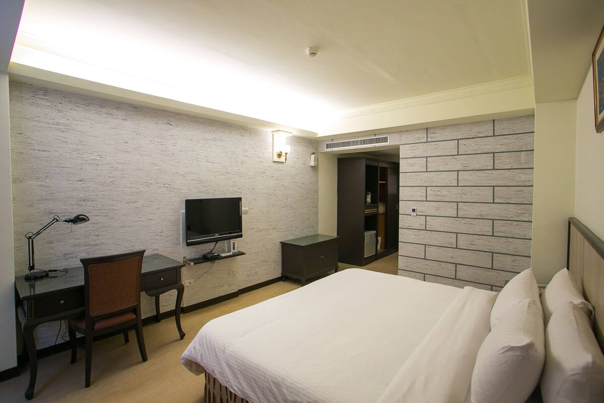 A hotel room with a bed, desk, chair, television, and a wardrobe.