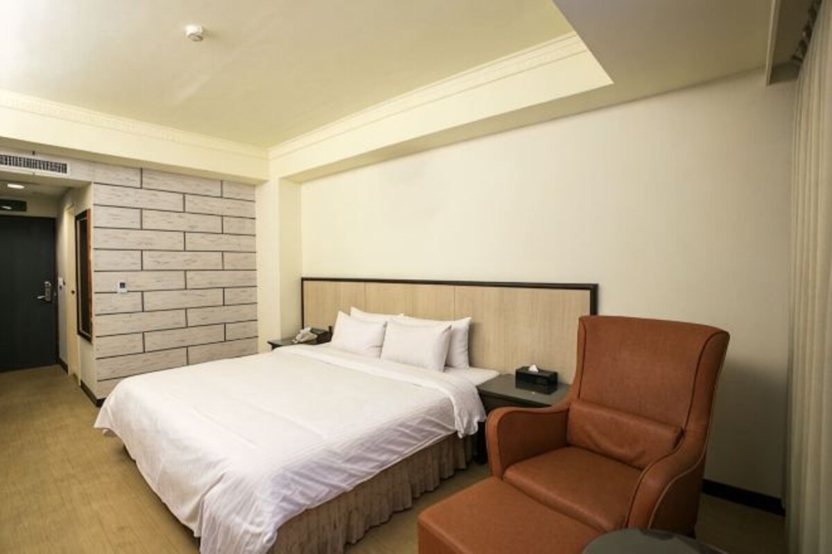 A hotel room with a bed, a nightstand, a chair, a brick-patterned wall, and a door.