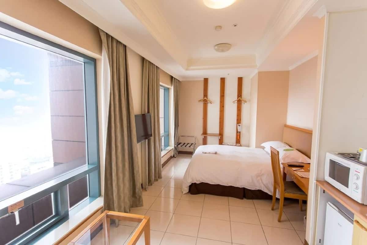 A hotel room with a large window, a bed, a desk, a chair, a TV, and a microwave.