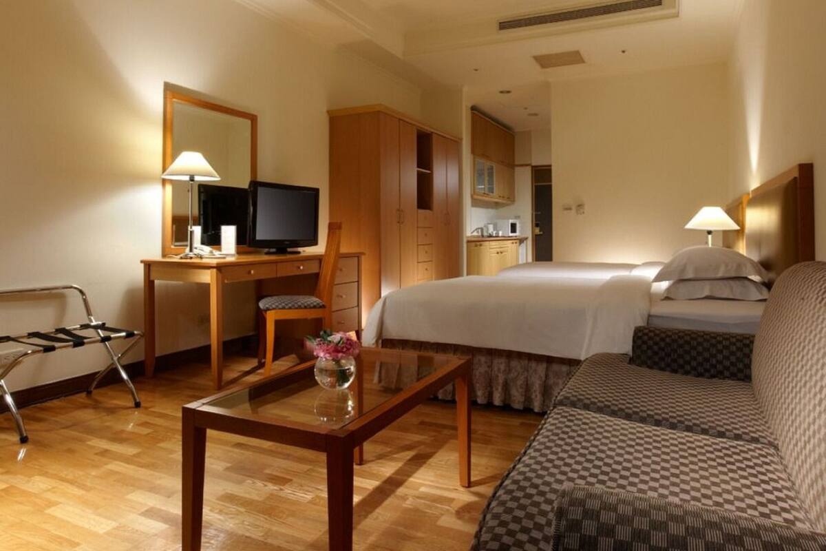 A hotel room with a bed, a sofa, a desk with a TV, a chair, and a small table with a vase.