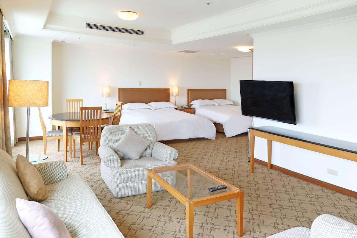 A hotel room with a sofa, armchair, two beds, a TV, and a small table.