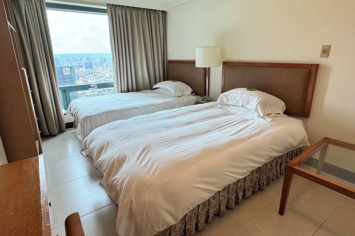 A hotel room with two beds, a desk, a lamp, and a view of the city.