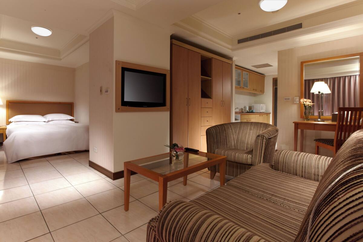 A hotel room with a bed, a TV, a sofa, a coffee table, a chair, and a kitchenette.