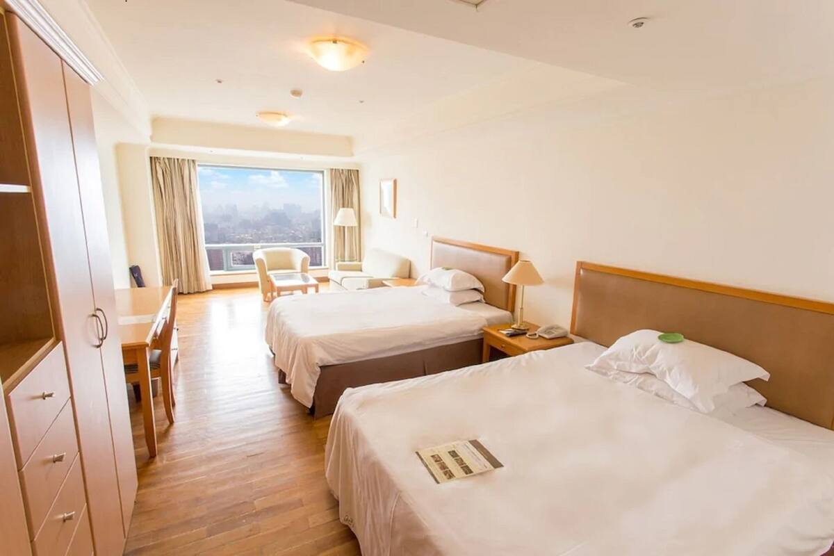 A hotel room with two beds, a desk, and a chair. There is a window with a view of the city.