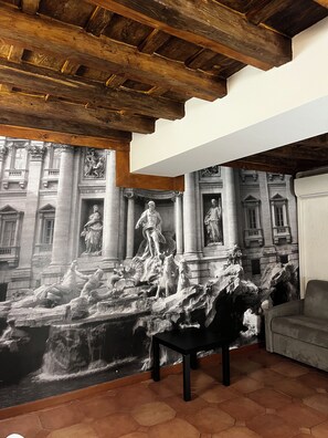 Une pièce ornée d’une grande fresque représentant des éléments d’architecture classique et d’une fontaine, avec un plafond en bois et un canapé.