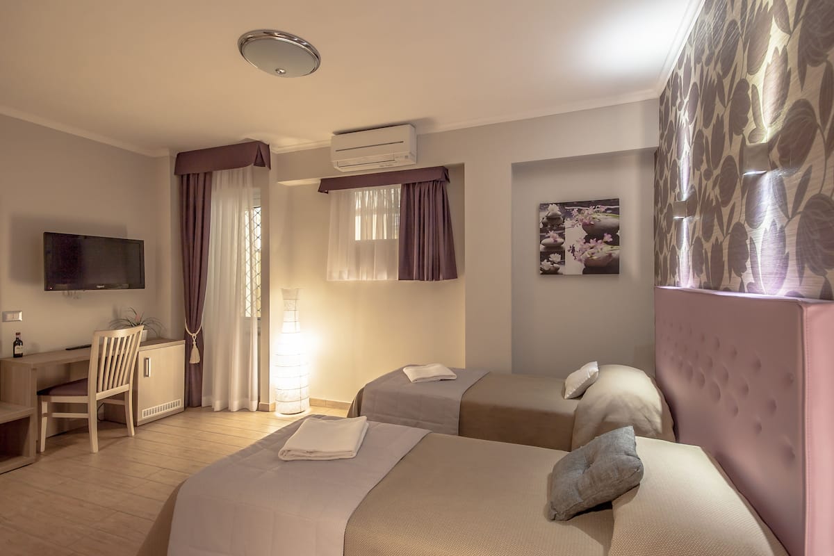 A hotel room with two beds, a desk, a chair, a TV, and an air conditioning unit.
