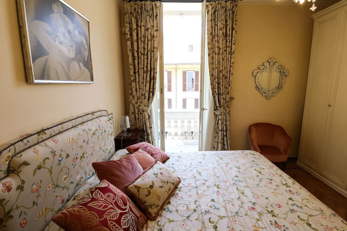 A bedroom with a bed, a chair, a painting, and a view of the outside.