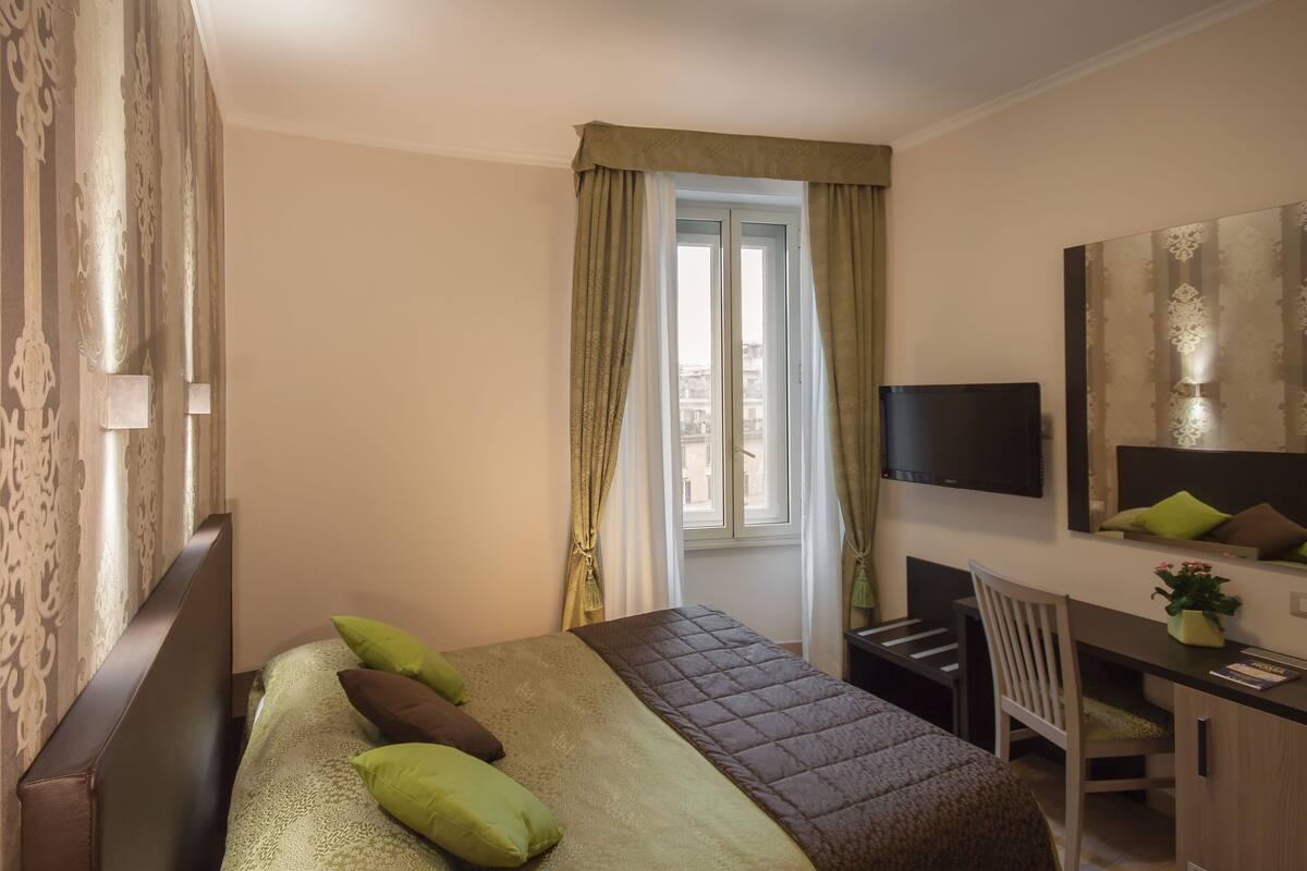 A hotel room with a bed, a desk, a chair, a TV, and a window with curtains.