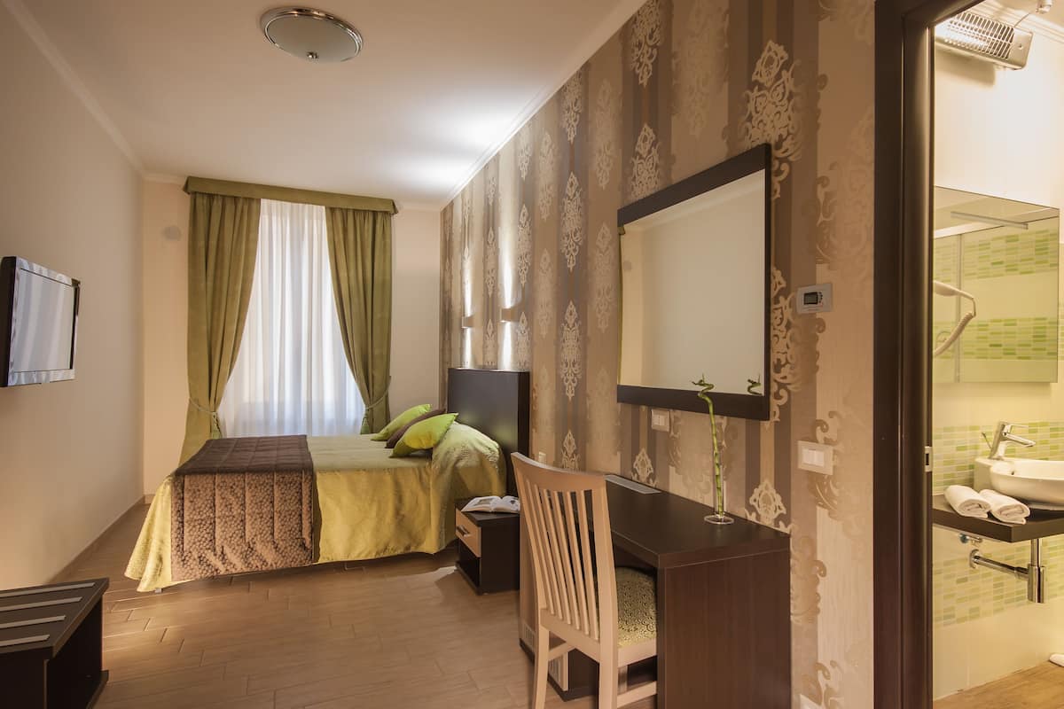 A bedroom with a bed, a desk, a chair, a TV, and a bathroom with a sink and mirror.