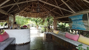 A covered outdoor seating area with a thatched roof, a stone bench, and a mural on the wall. - Flamingo Villas Resort (Malindi)