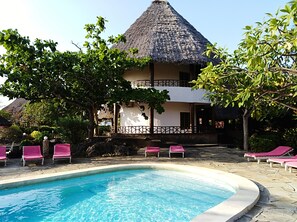 A pool area with lounge chairs, a thatched-roof building, and lush greenery. - Flamingo Villas Resort (Malindi)