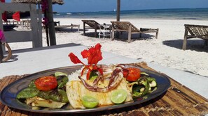 A beachside dining table with a plate of grilled vegetables and rice, surrounded by beach chairs and a thatched umbrella. - Flamingo Villas Resort (Malindi)