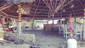 An outdoor seating area with thatched roofs, stone walls, and a grill sign in the background. - Flamingo Villas Resort (Malindi)