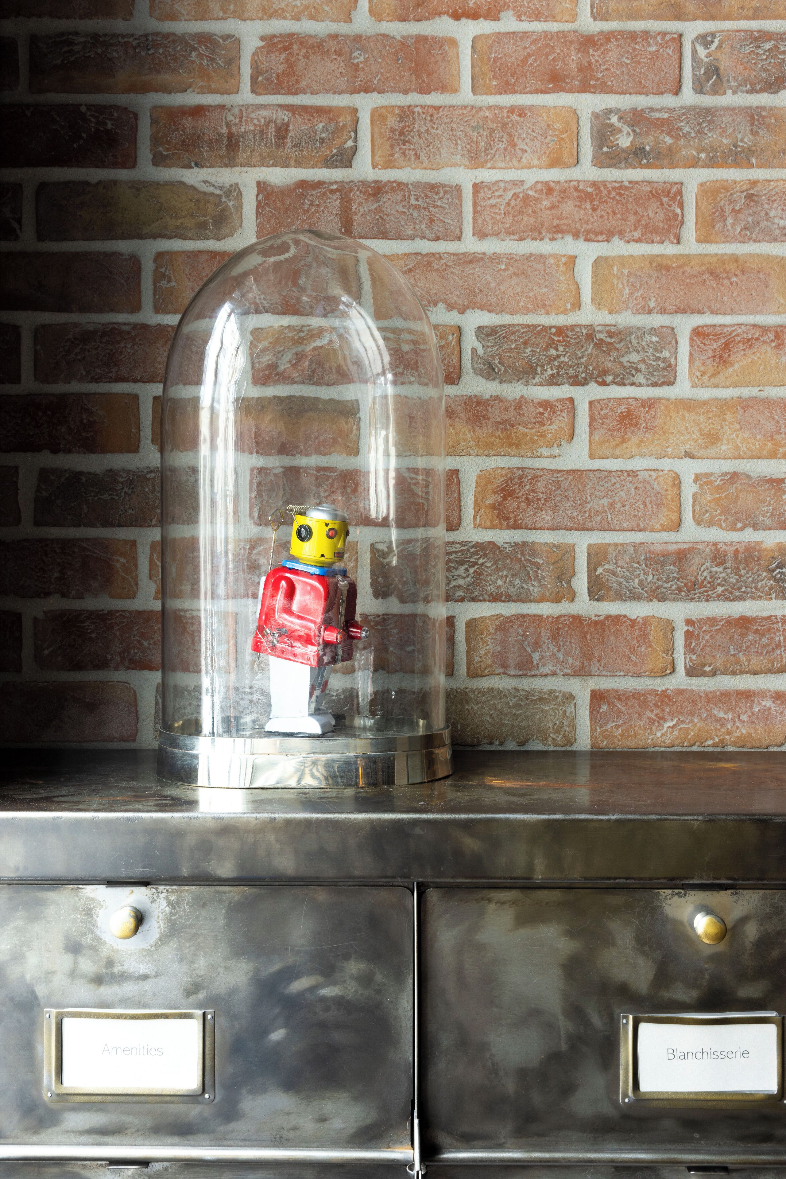 Un robot jouet, placé sous une cloche de verre sur une surface métallique, avec des tiroirs étiquetés « Fournitures » et « Laverie ».