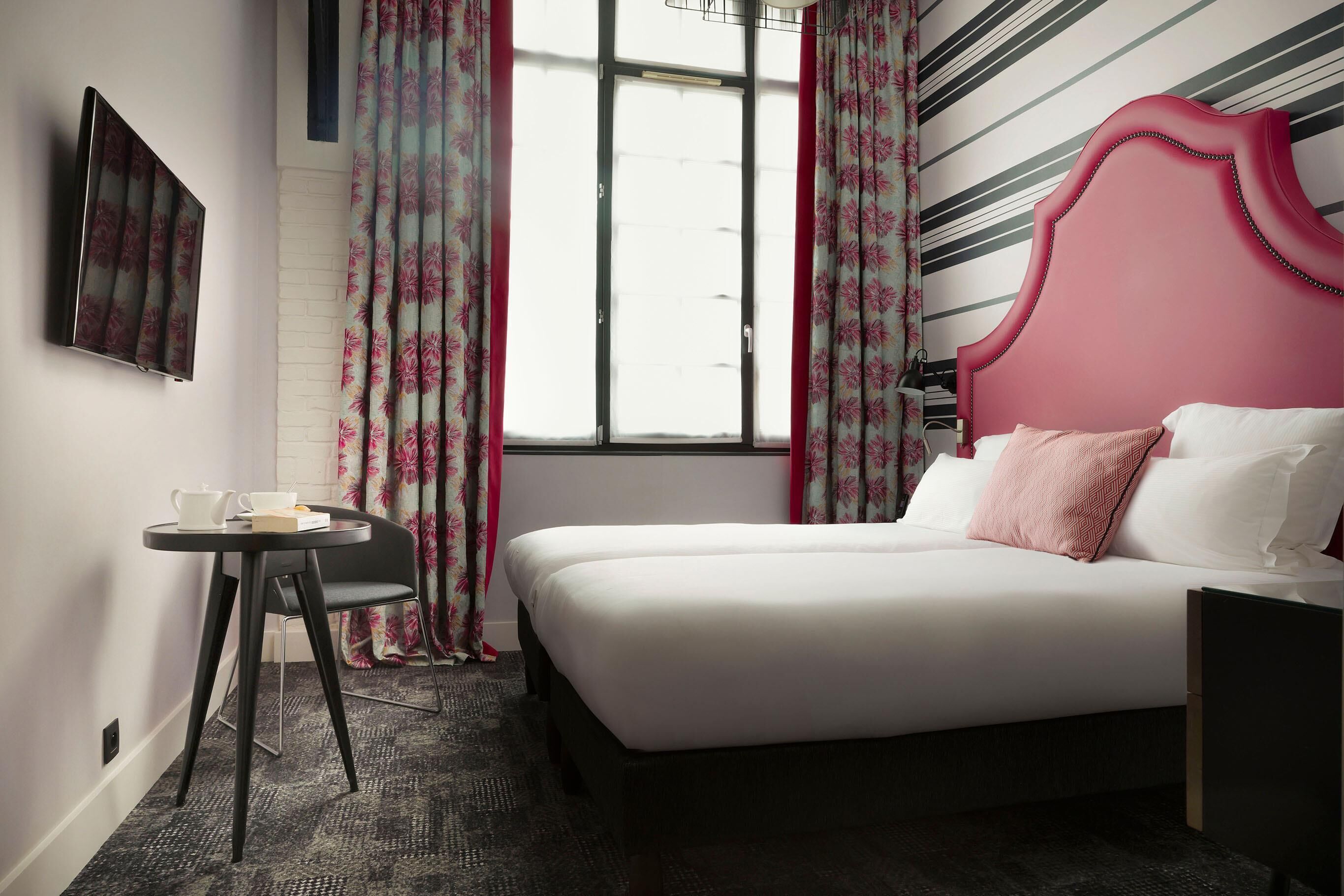 Una camera d'albergo con un letto, un piccolo tavolo con una tazza e un libro, e tende a motivi floreali.