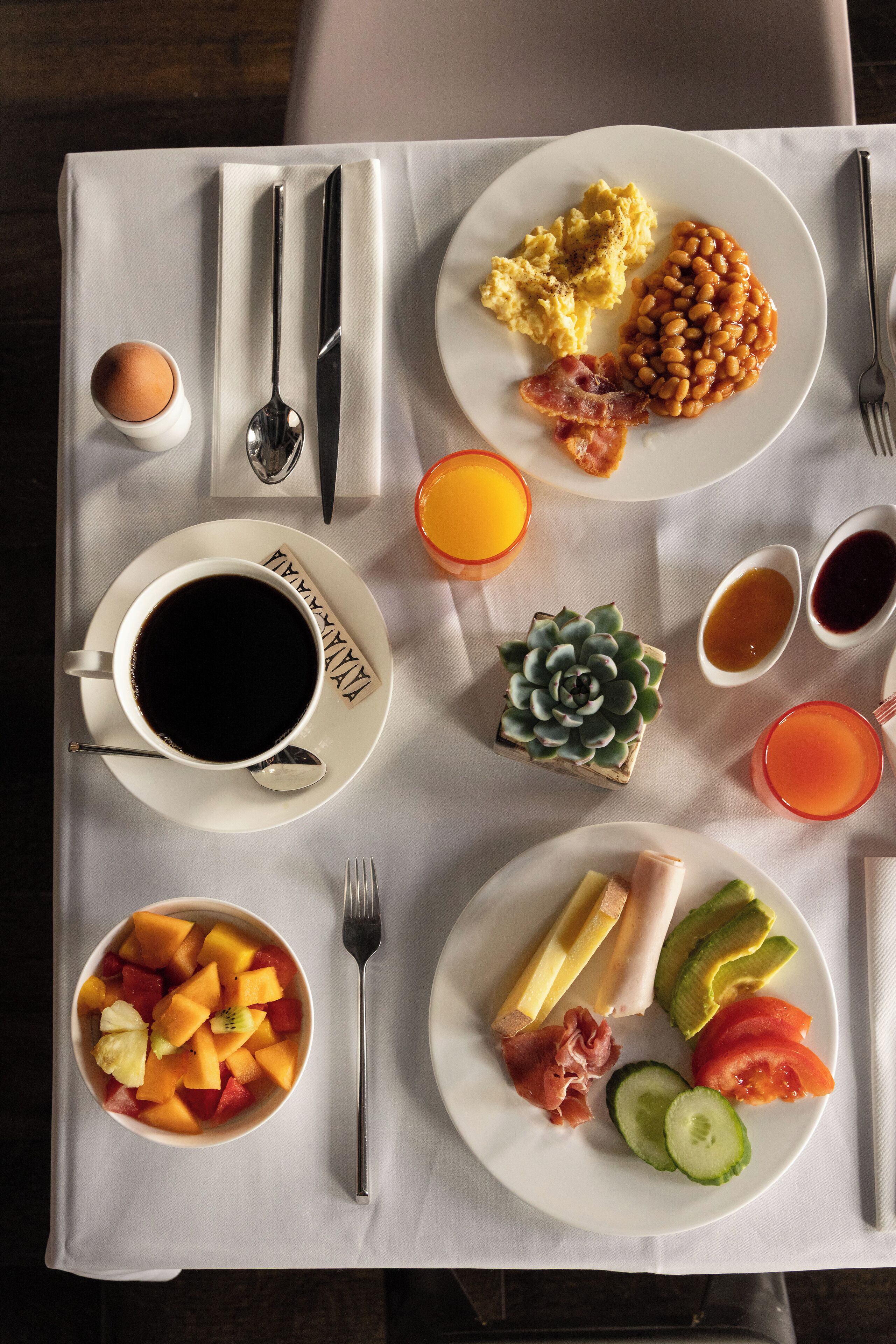 Ein Frühstückstisch mit Tellern voller Eier, Bohnen, Speck und Obst, sowie Tassen Kaffee und Saft.