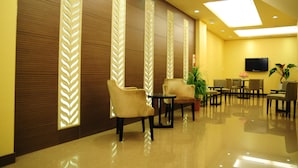 Un vestíbulo de hotel con piso pulido, zona de estar con sillas y mesas, paneles decorativos en la pared y un televisor de pantalla plana montado.