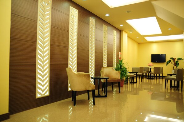 Um hall de hotel com piso polido, área de estar com cadeiras e mesas, painéis decorativos na parede e uma TV de tela plana fixada na parede.