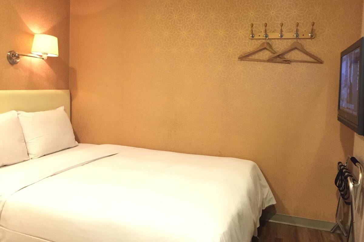 A hotel room with a bed, a wall-mounted light, and a coat rack.