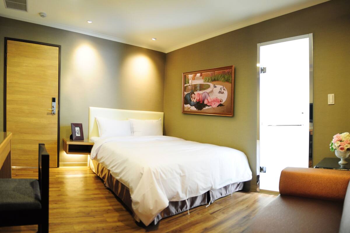 A hotel room with a bed, bedside table, nightstand, bench, and a door.