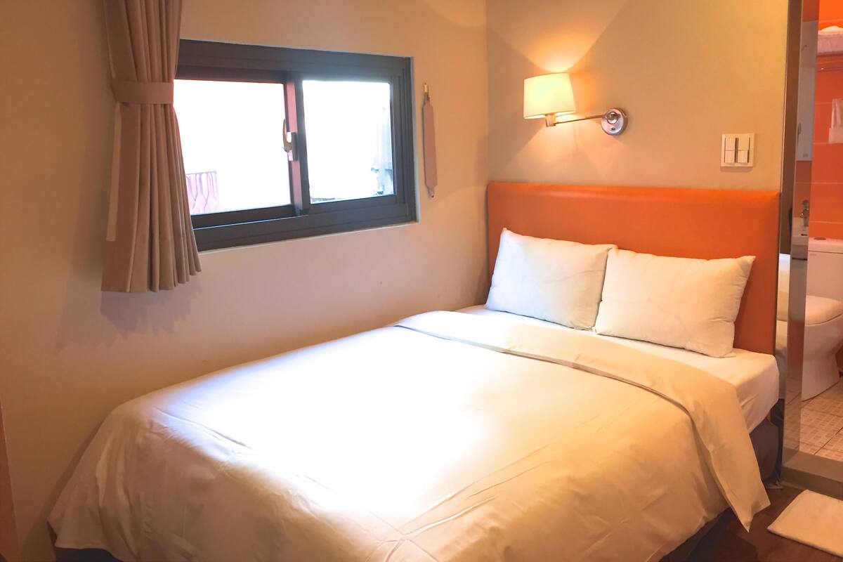 A hotel room with a bed, a window with curtains, a wall-mounted lamp, and a visible bathroom.