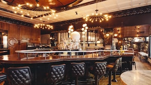 A bar area with wooden bar counters, black cushioned bar stools, and a variety of bottles and glasses.