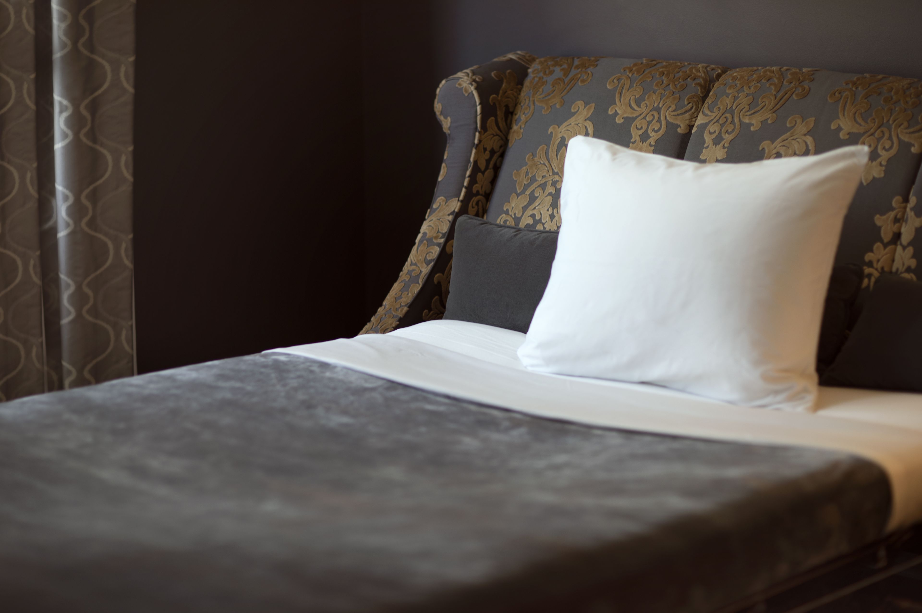 Un letto rifatto con un piumone grigio e un cuscino bianco, una testiera scura con motivo decorativo e una tenda con motivo ondulato.
