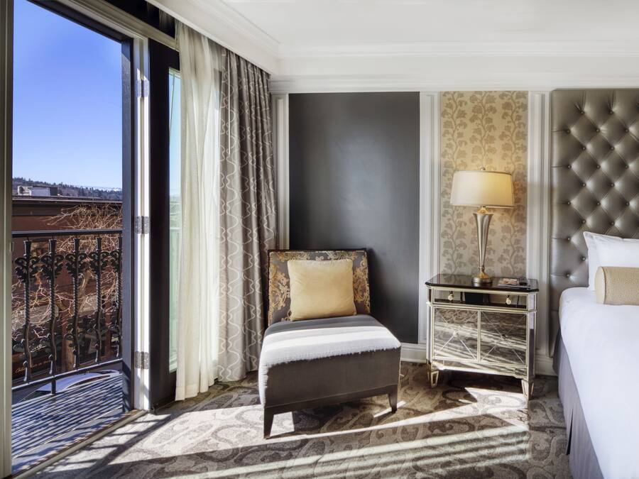 A hotel room with a bed, a chair, a nightstand, a lamp, and a view of the outdoors.