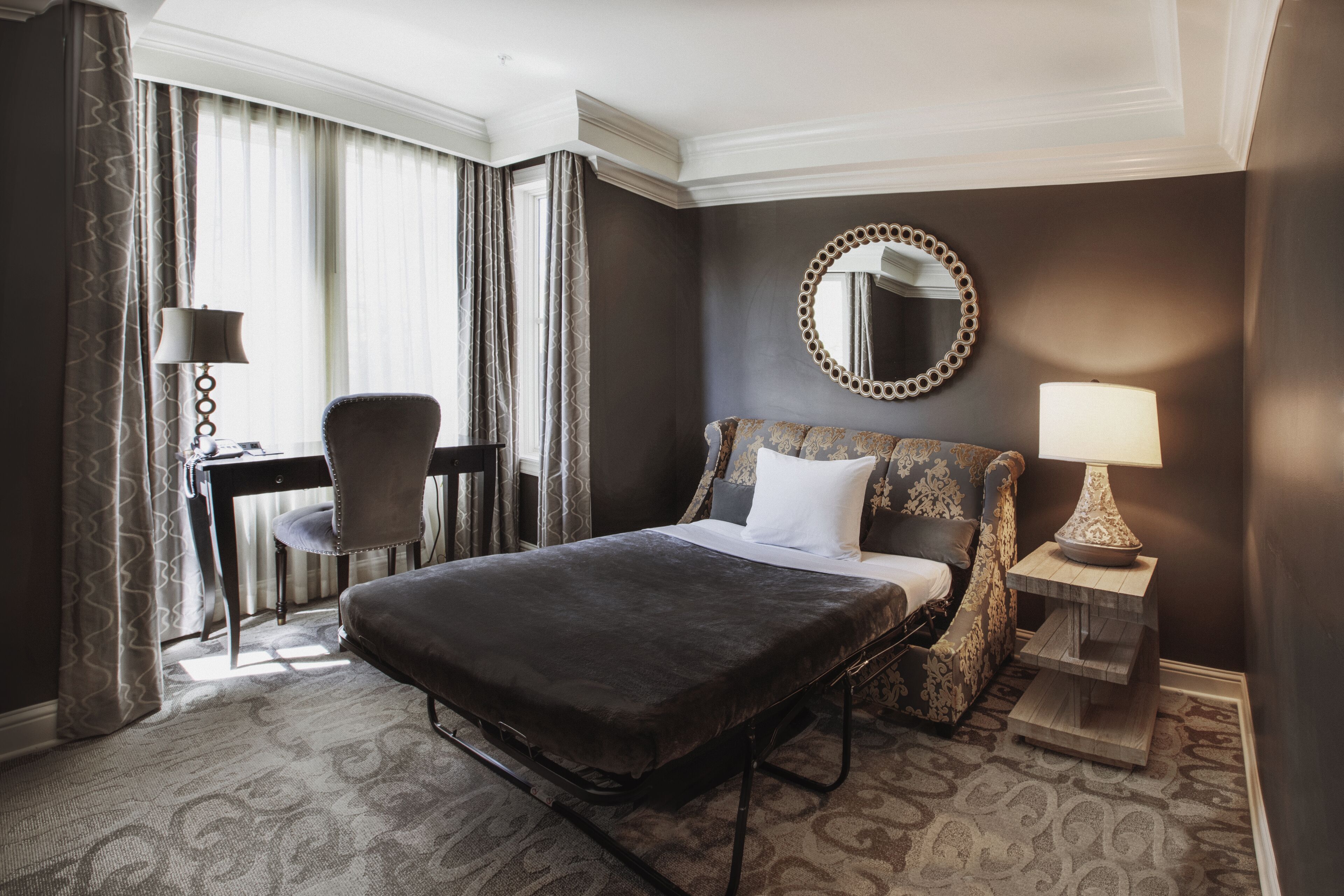 A hotel room with a large bed, a desk, a chair, a round mirror, and a lamp.