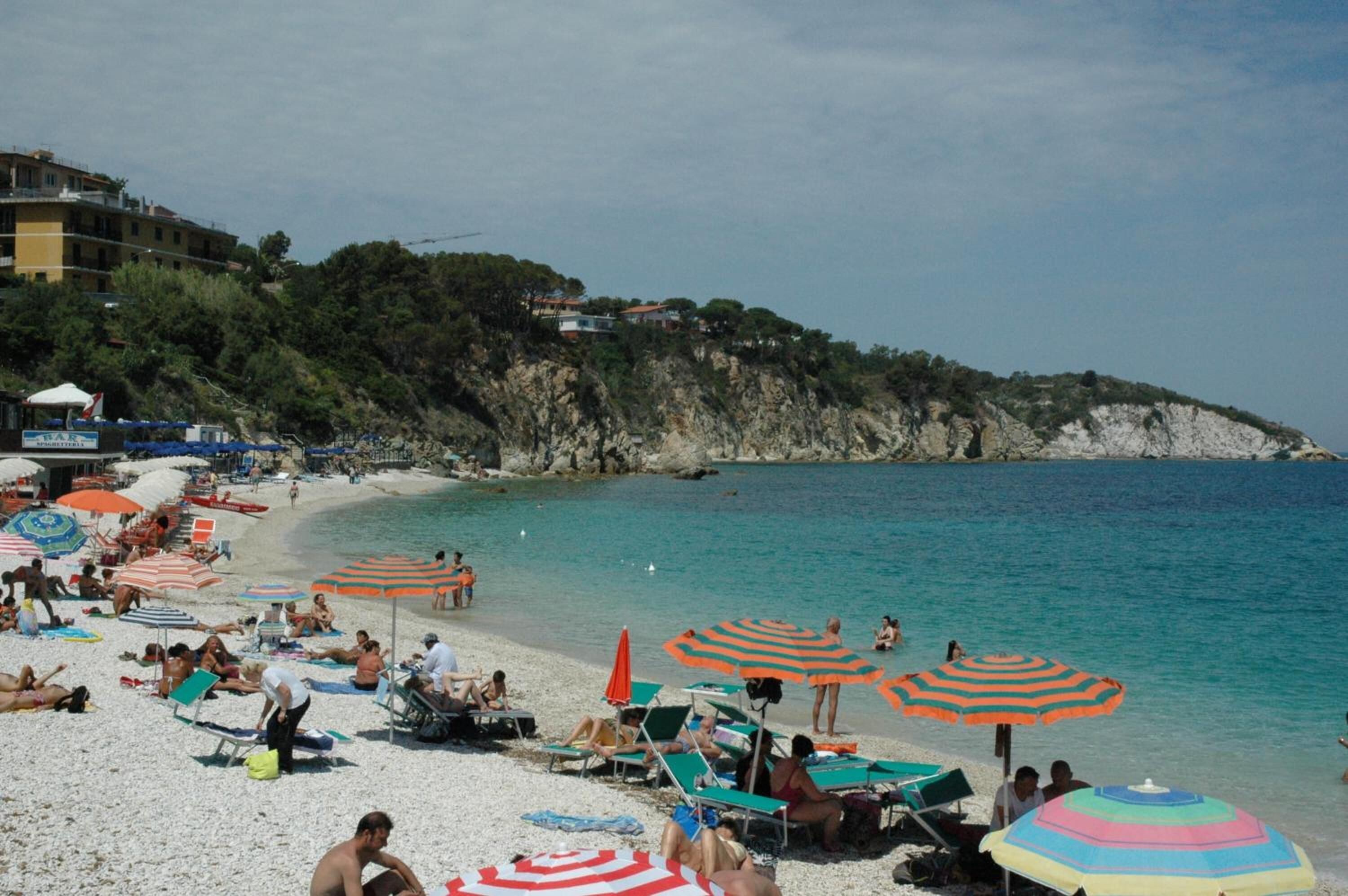 Une plage avec de nombreux parasols colorés et des chaises longues, une côte rocheuse et des bâtiments en arrière-plan.