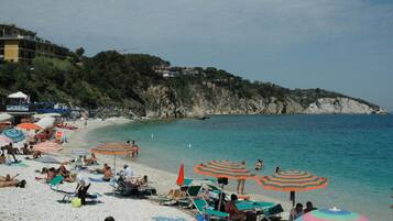 Une plage avec de nombreux parasols colorés et des chaises longues, une côte rocheuse et des bâtiments en arrière-plan.