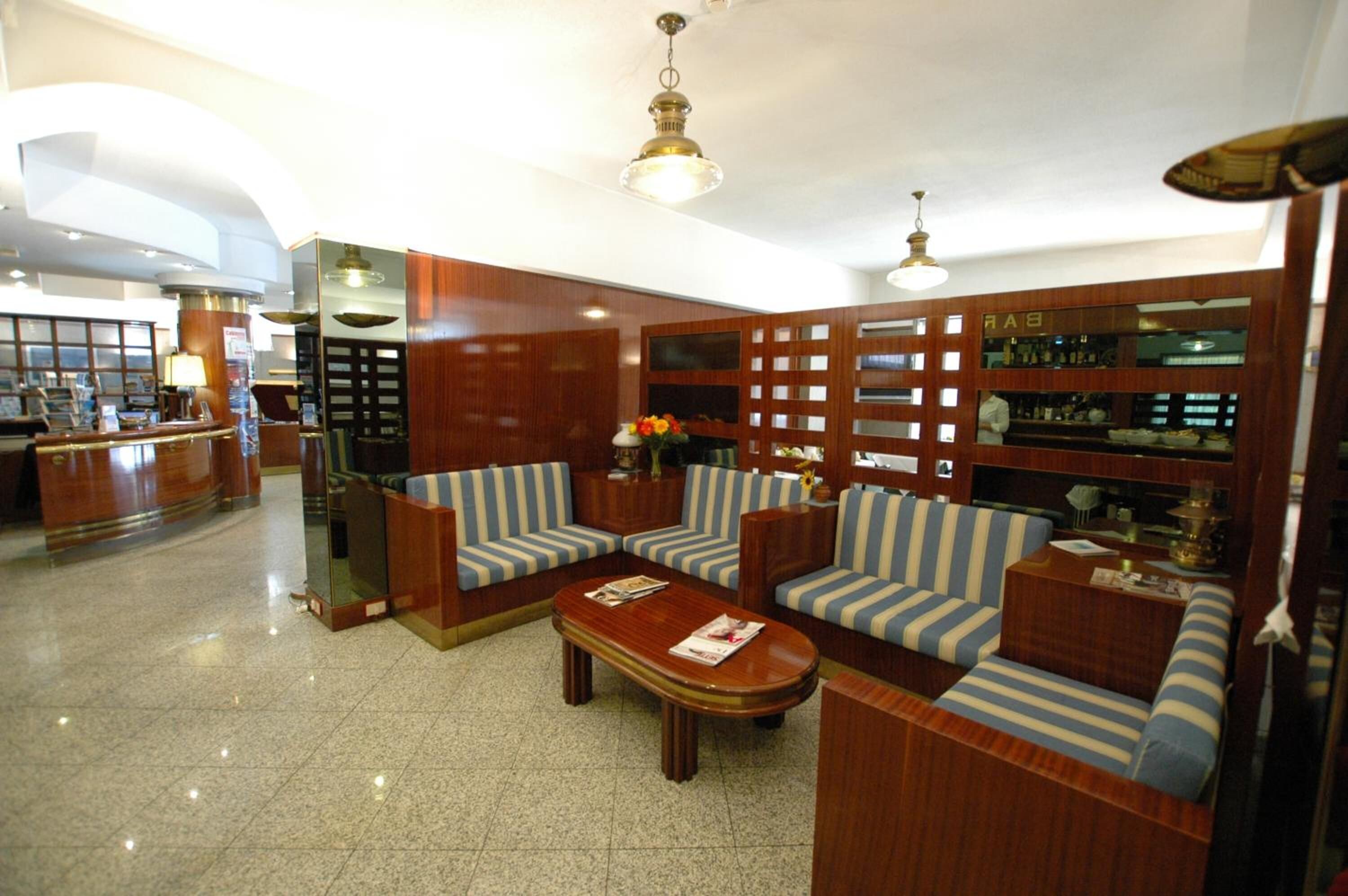 A lobby with a reception desk, striped sofas, a wooden coffee table, and a bar area with a sign reading "BAR".