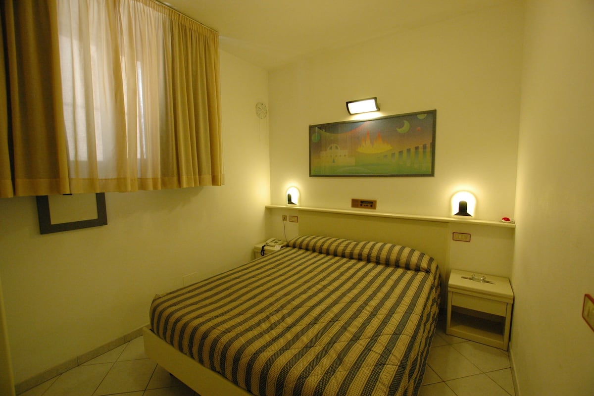A hotel room with a bed, a nightstand, a lamp, a painting on the wall, and a window with curtains.