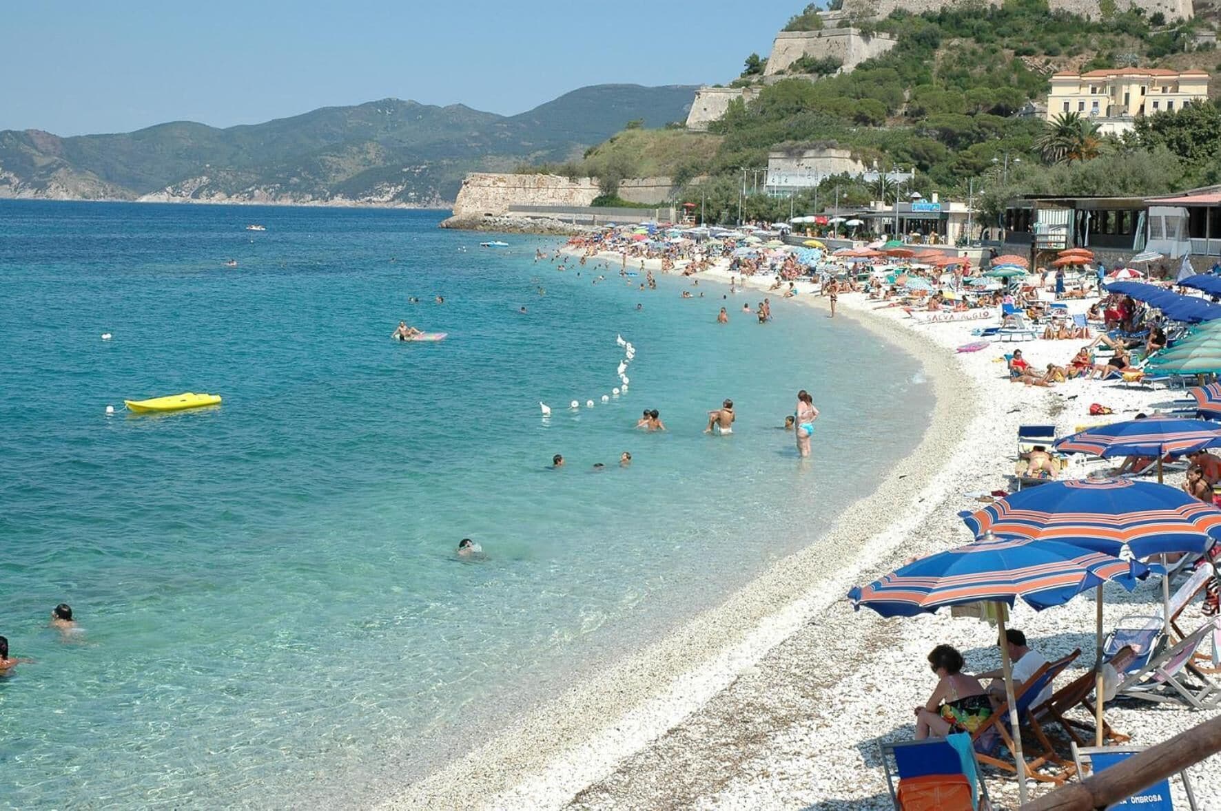 A crowded beach with clear blue water, umbrellas, and people swimming.