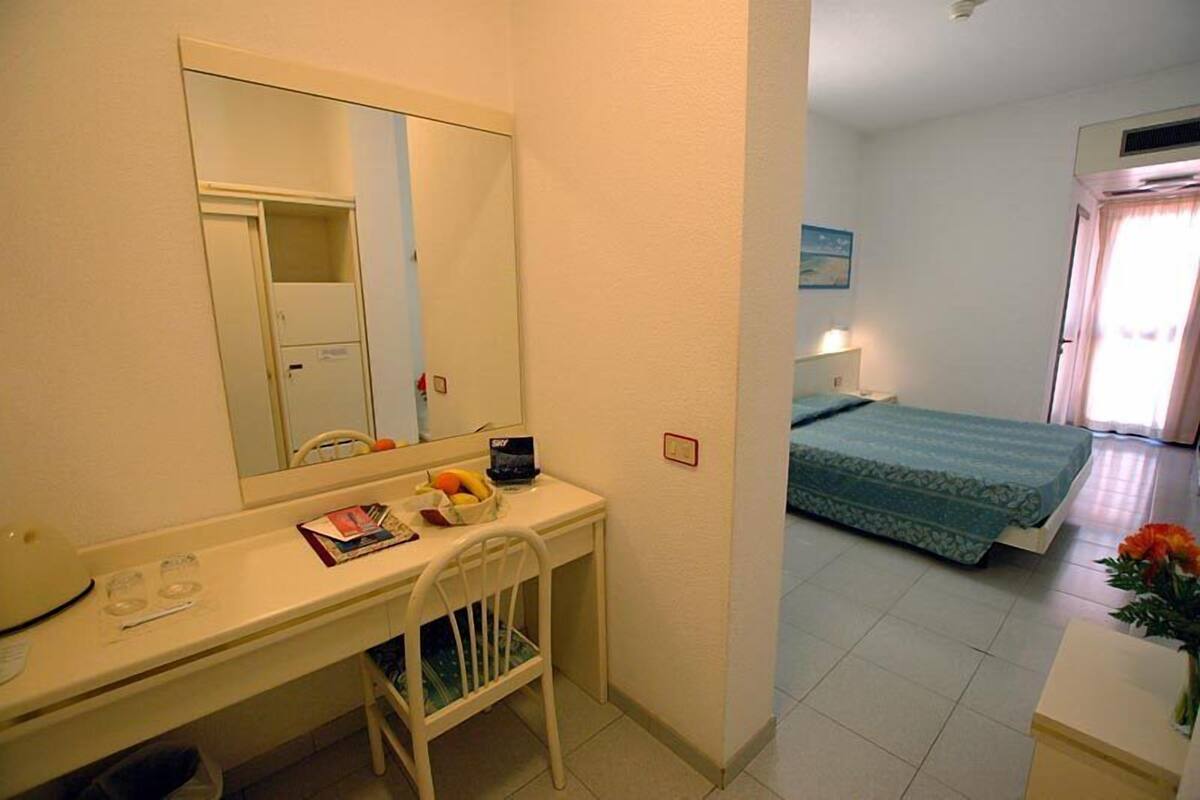 A hotel room with a bed, a desk with a chair, a mirror, and a small table with a vase of flowers.