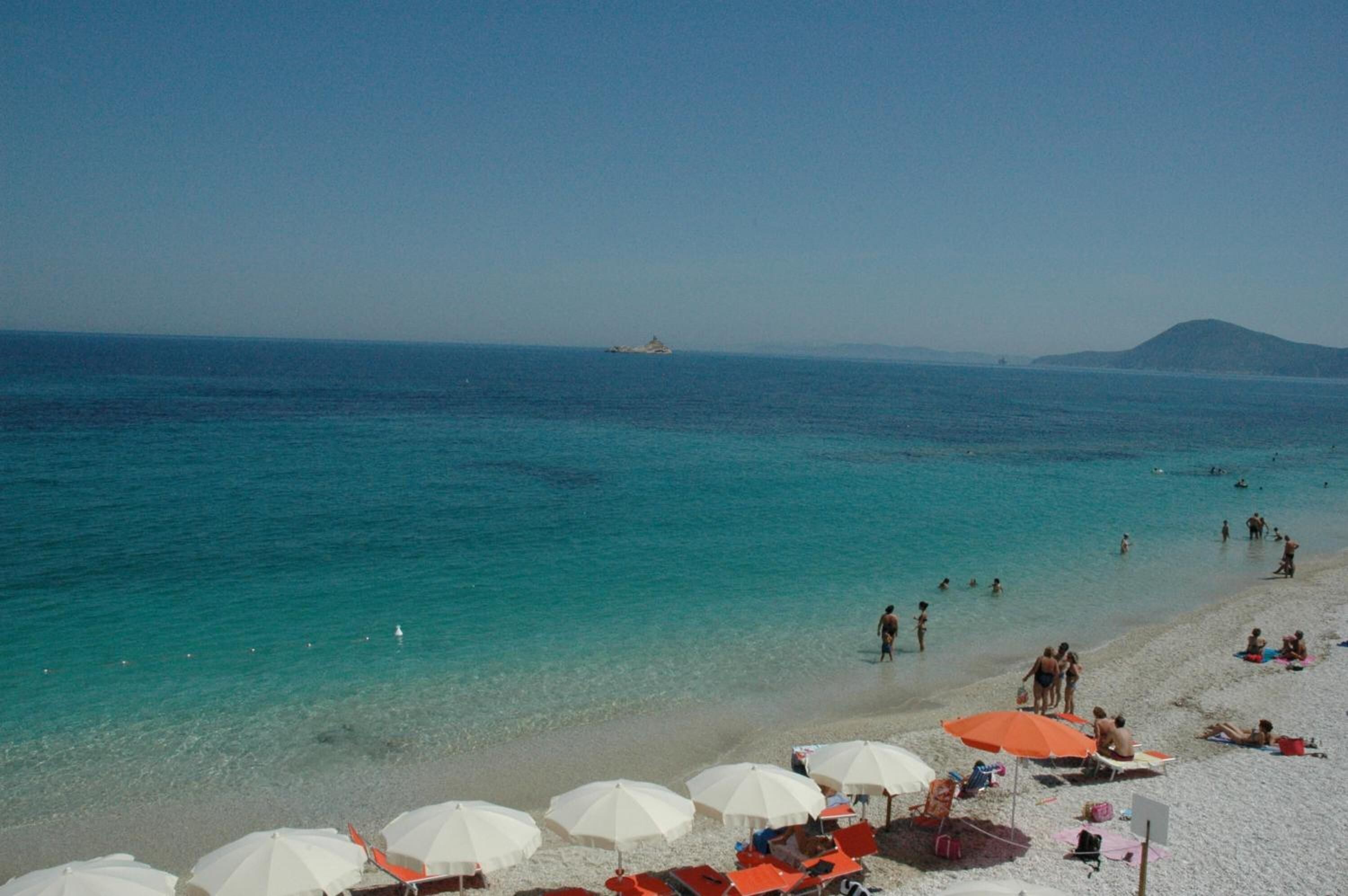 Ein Strand mit klarem türkisblauem Wasser, weißen Sonnenschirmen und Menschen, die sich sonnen.