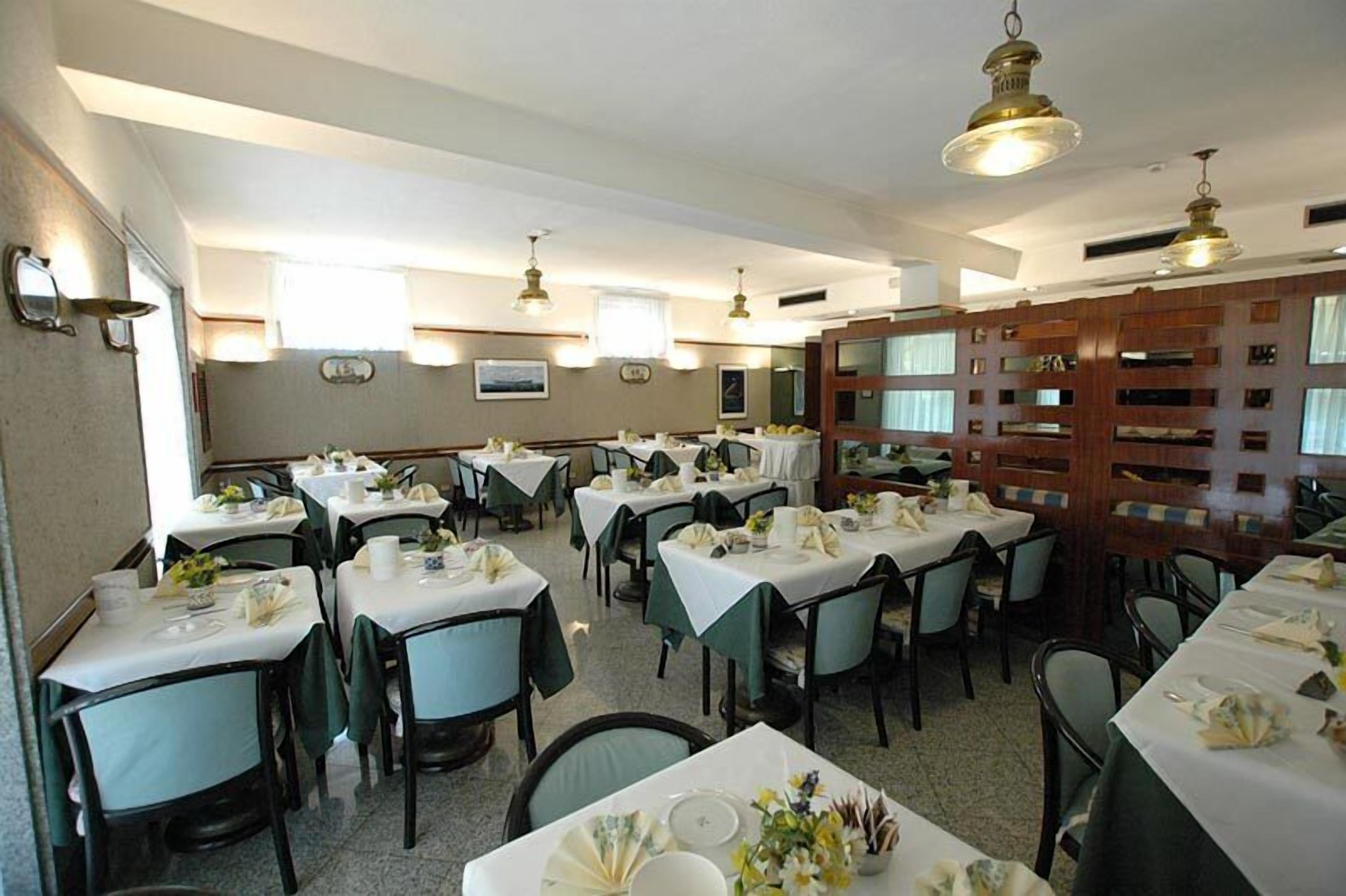 Une salle à manger avec plusieurs tables dressées pour le service, chacune étant recouverte d’une nappe blanche et entourée de chaises vertes.