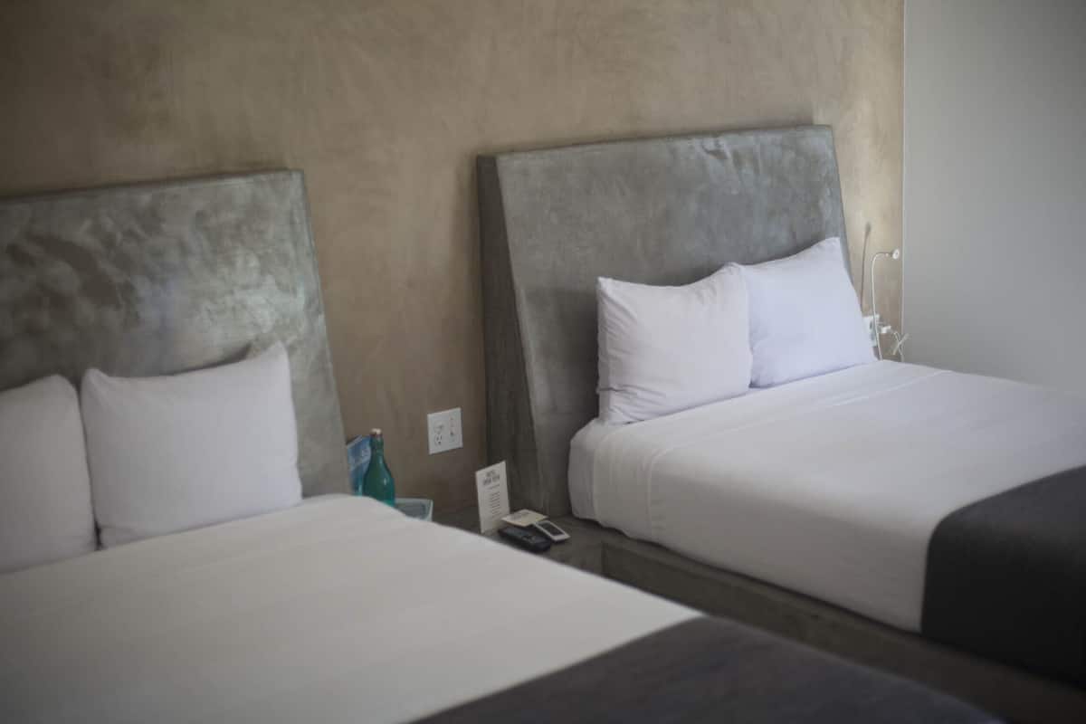 Dos camas con cabeceros grises, ropa de cama blanca, una mesita con una botella y un control remoto, y un enchufe de pared.
