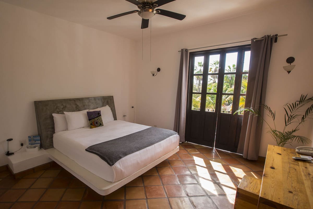 Un dormitorio con cama, ventilador de techo, ventana con cortinas, mesita de noche y un pequeño mueble de madera junto a la cama.