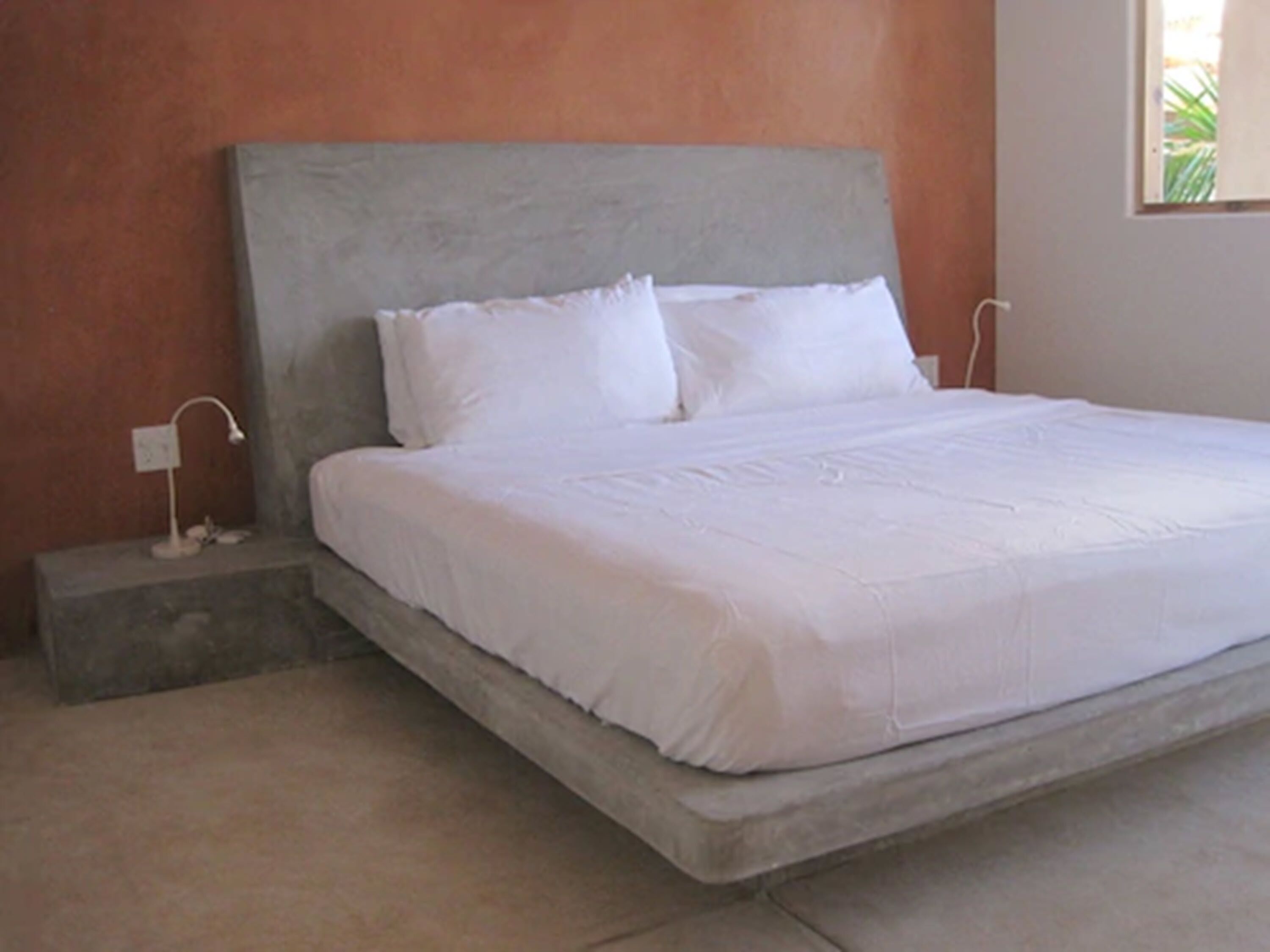 A large bed with white linens in a room with a plain wall and a small bedside table with a lamp.