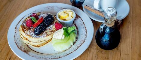 A plate of pancakes with berries and a small bowl of fruit, a cup of coffee, and a bottle of syrup on a wooden table.
