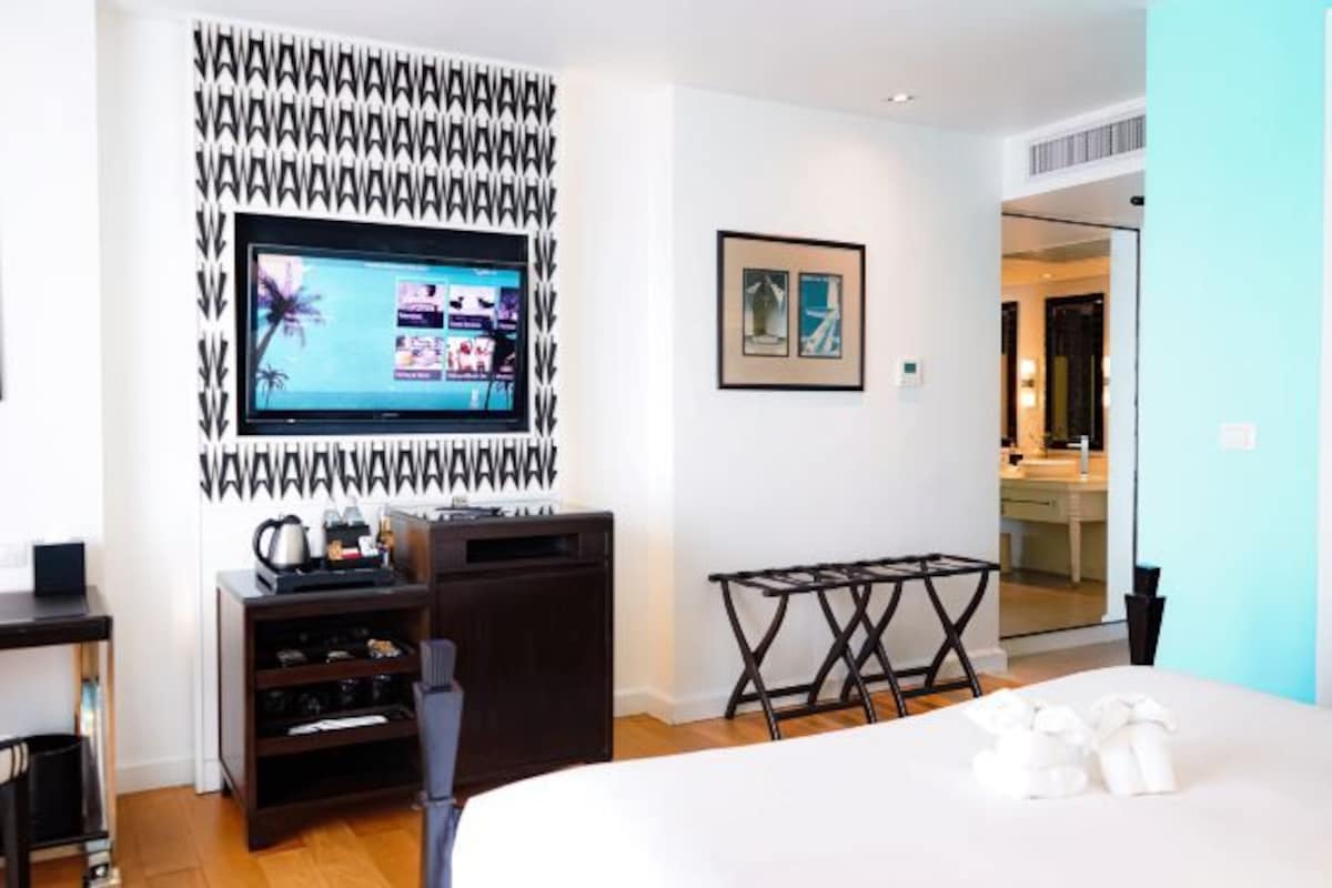 A modern hotel room with a flat-screen TV mounted on the wall, a small wooden side table, a bed with neatly folded towels, and a framed picture on the wall.