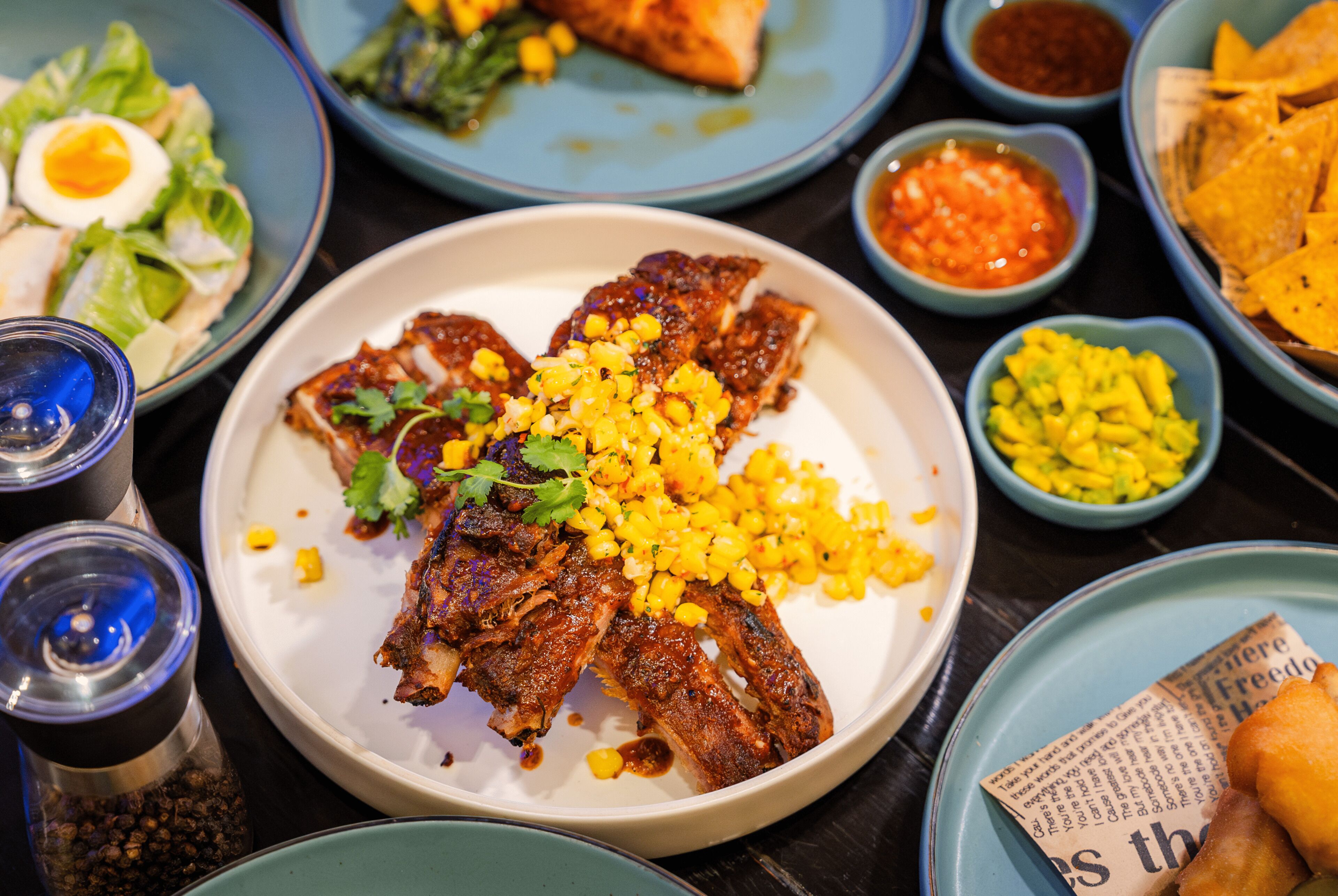 A variety of dishes including a plate of ribs with corn and greens, a bowl of corn salad, and several small bowls of sauces.