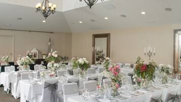 A banquet hall with multiple tables set for a formal event, featuring chandeliers, floral centerpieces, and a high ceiling with skylights.