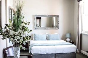 Quarto com cama, espelho, um vaso de flores e abajur em uma mesinha de cabeceira.