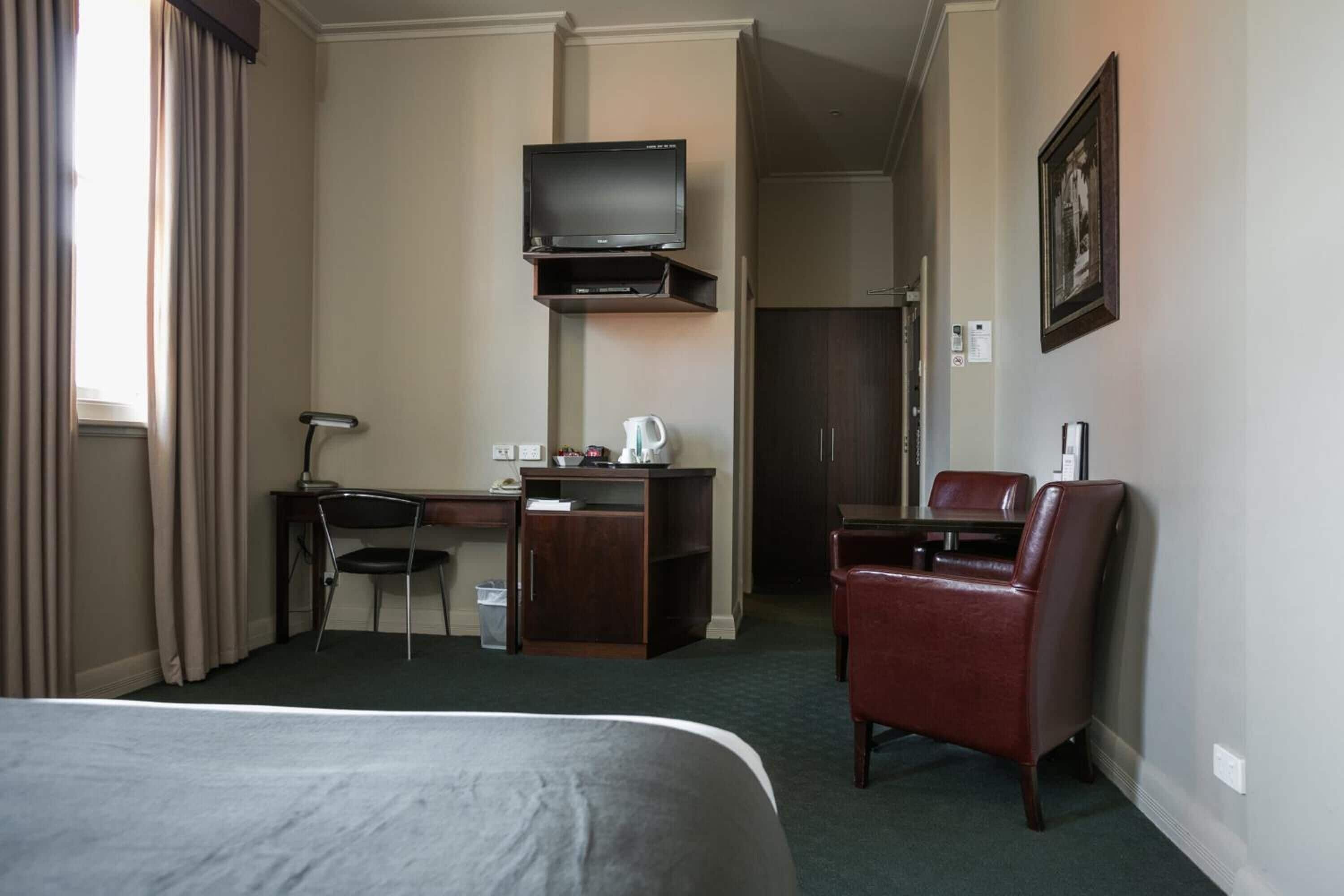 A hotel room with a bed, desk, chair, TV, and a small kitchenette.