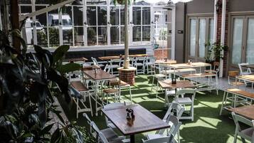 An outdoor cafe with tables and chairs arranged on artificial grass, surrounded by large windows and potted plants.