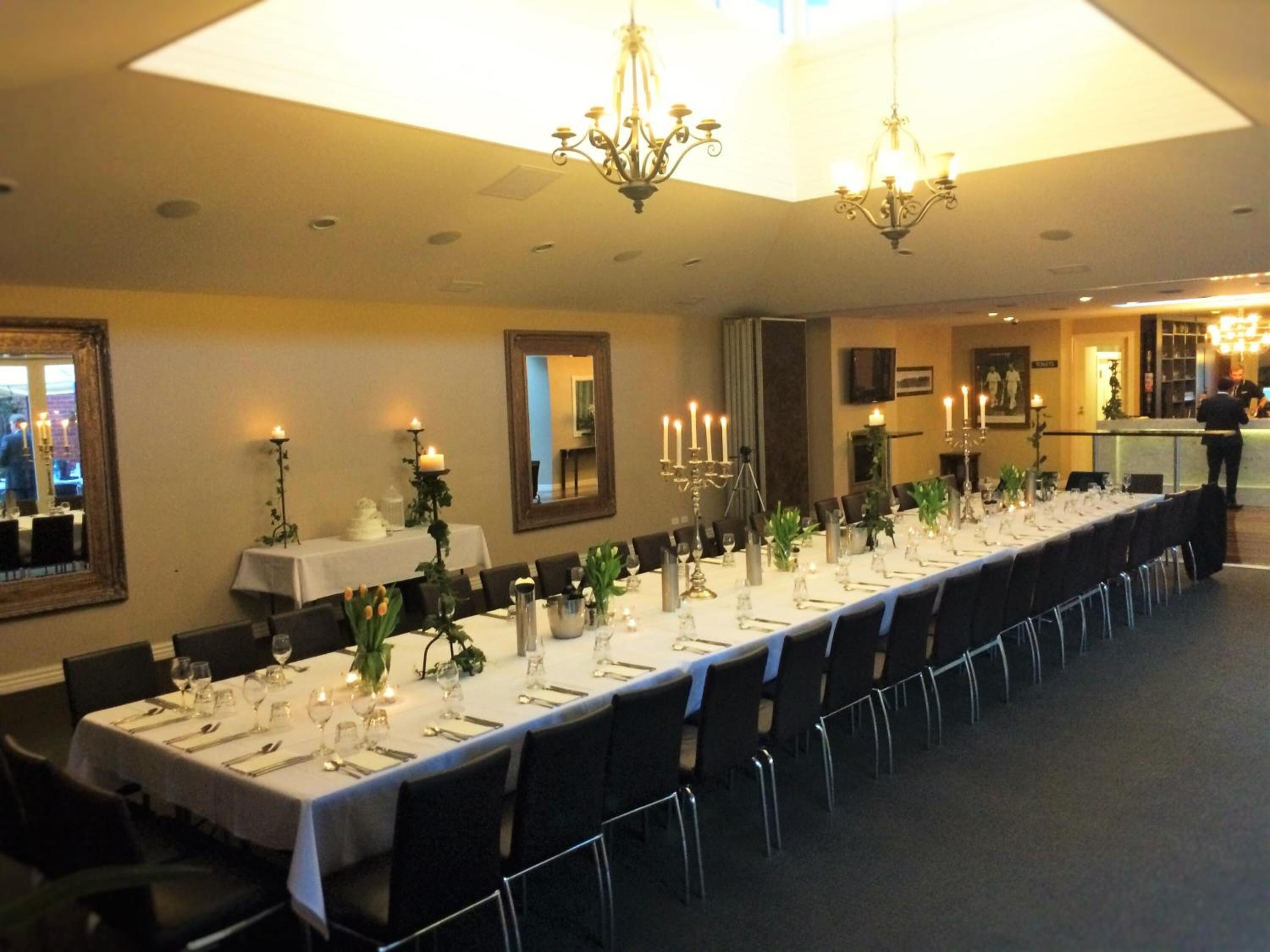 A long dining table set with white tablecloths, candles, and glassware, in a room with chandeliers and framed artwork on the walls.